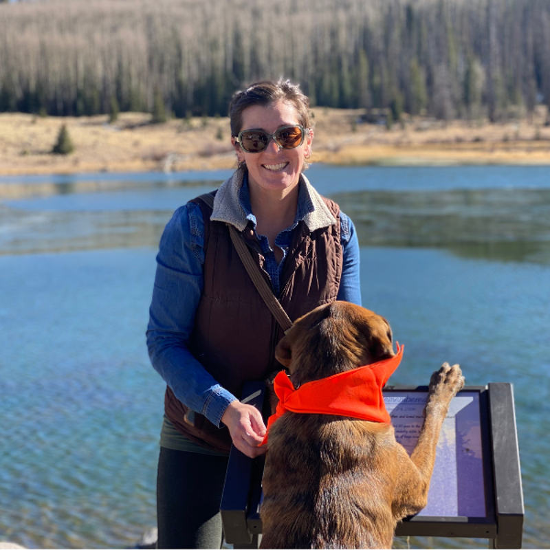 A woman and a dog are standing next to a lake. The woman is wearing sunglasses, a denim shirt, and a brown vest. The dog is wearing an orange bandana and is standing on its hind legs, with its front paws resting on a sign. The lake is blue and there are trees in the background. The sky is clear and sunny.